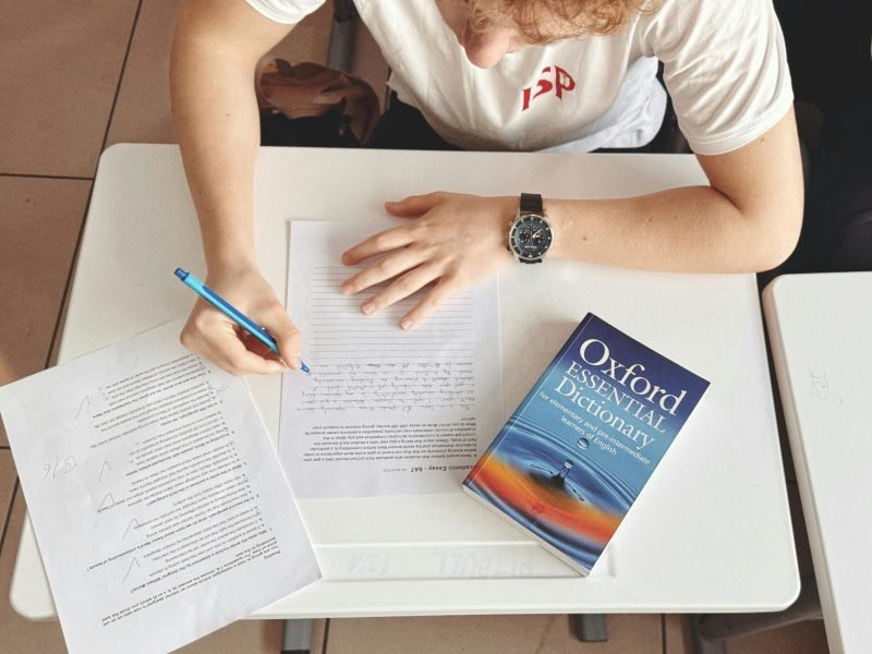 Student writing test using paper and Oxford dictionary on desk in classroom